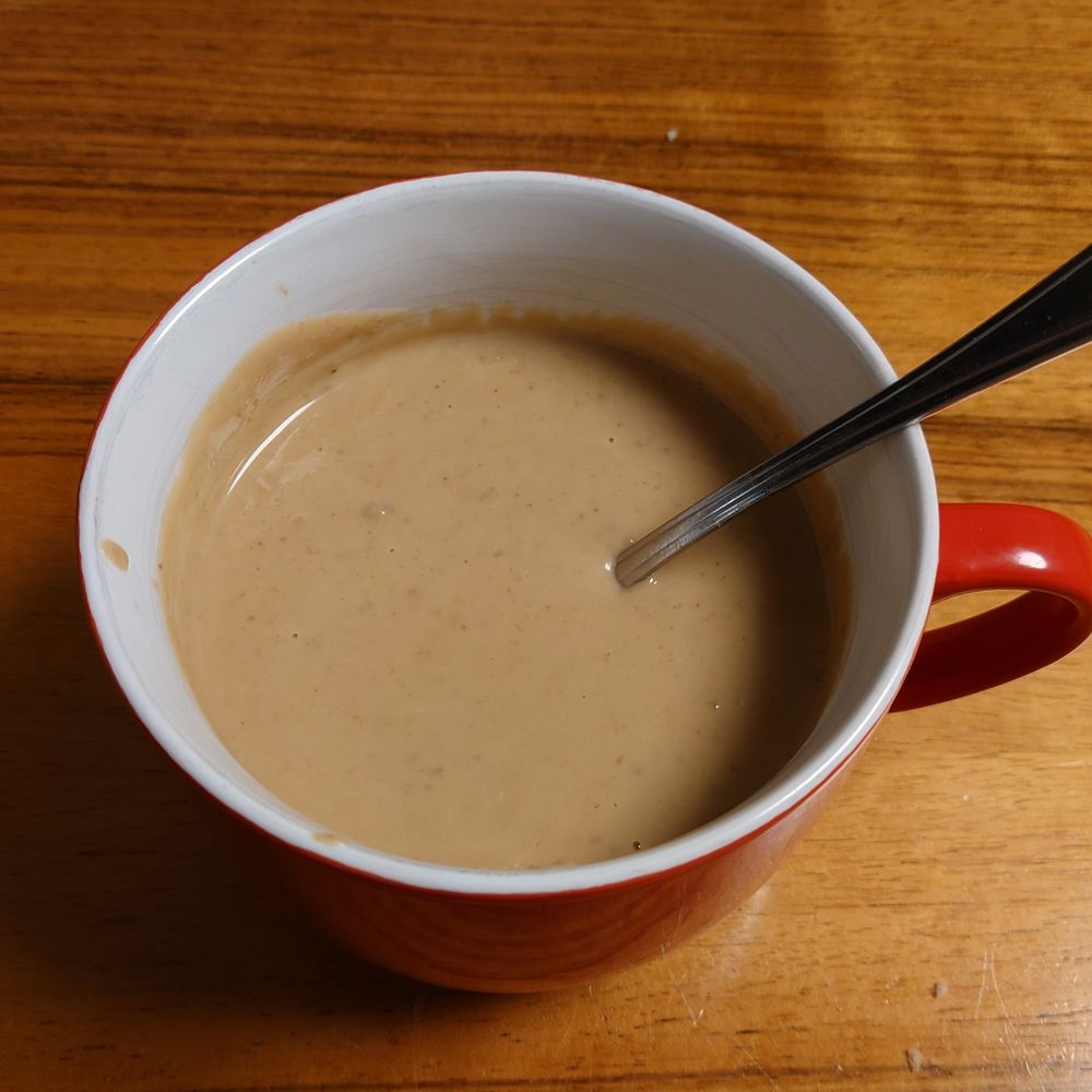 Auf einem Holztisch steht eine große, rote Tasse mit Henkel in der hellbraune Maronensuppe ist und ein Löffel guckt raus.