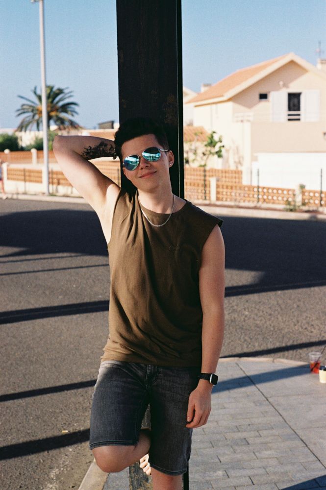 A young man with dark hair leans casually against a dark post outdoors, smiling at the camera. He wears round, reflective sunglasses, an olive green sleeveless t-shirt, and dark shorts. His right arm is bent behind his head, revealing a tattoo on his upper arm. The background shows a road, a house with a terracotta roof, and a palm tree under a bright blue sky. The image has the warm, grainy quality of a film photograph.