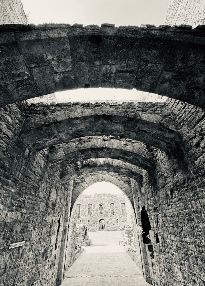 A silver-toned black and white photograph of the entrance to the outer ward of a stone castle. Overhead, the gaps between stone arches serve as murder holes.