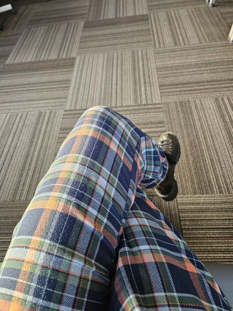 Picture of a person's legs crossed sitting in a chair wearing navy blue, peach, & green plaid pants and lace up black ankle boots. 