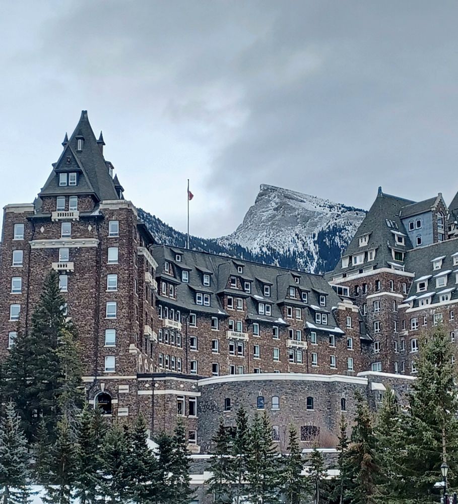 A photo of Fairmont Banff Springs Hotel, a tall castle made of brown bricks. It's framed by pine and the rockies peek over it's spike rooves, cut out of a grey sky.