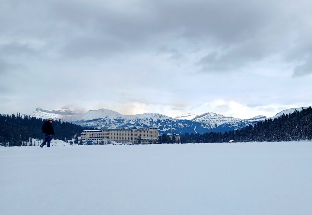 A low angle photo from the middle of a frozen over Lake Louise, of the mountain line behind Chateau Lake Louise, cast in a subtle morning glow.