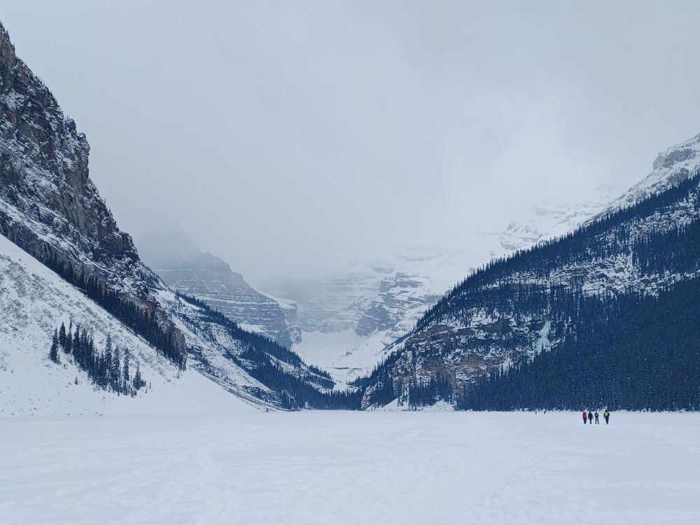 A photo of Lake Louise's blue tinted frozen mountains taken from the middle of a frozen over Lake Louise. The Victoria glacier is obscured by snow and clouds.