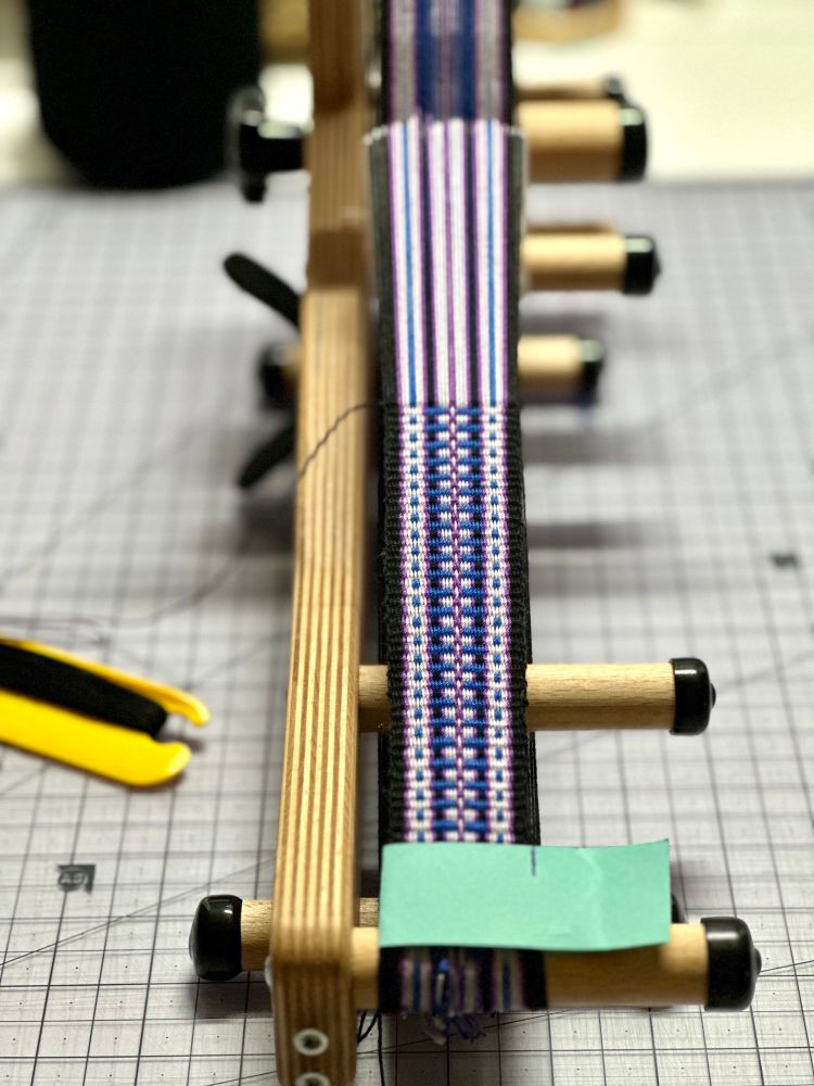 An inkle loom with a partial woven band sits on top of a checkerboard pattern. A yellow belt shuttle is on the left of the loom with black yarn wrapped around the center. 