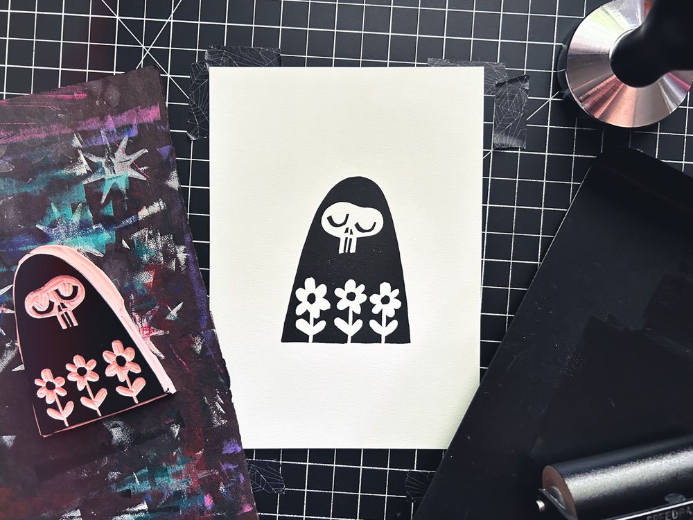 a black block print on white paper surrounded with block printing supplies. the print is of a hooded figure with a contented skull face looking at three flowers in negative in the foreground