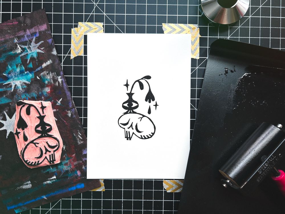 a block print surrounded by block printing supplies. the print is in black ink on white paper and depicts a sorry-looking skull with a vase with an eyeball on its head. in the vase is a drooping flower with single petal/raindrop falling from it.