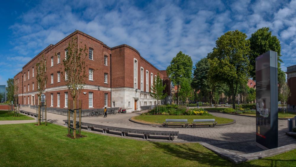A shot of Main Library from the side on, on a sunny day with blue skies, capturing the trees in front of the building and the benches