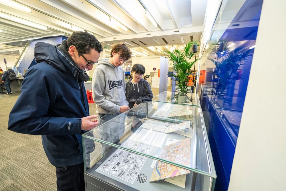 Three Library users looking at items on display in Main Library as part of the You Are Here! exhibition