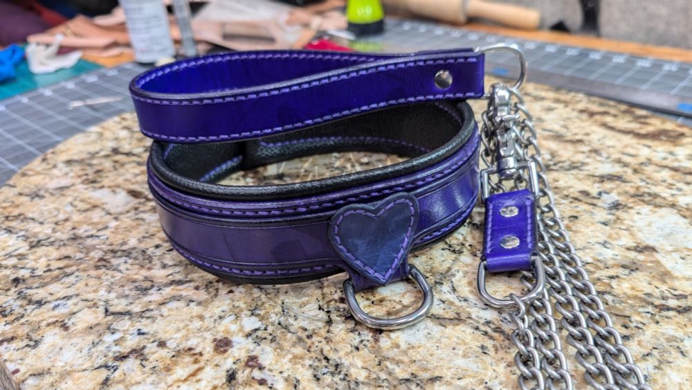 Purple and black leather collar with sewn-on lavender heart emblem and drop down D-ring, and a 4ft purple leather and steel leash. Purple top and back straps, black leather liner, light purple thread, stainless steel hardware with colored rivets to match. The leash has a purple leather handle and extender,, light purple thread, steel chain, and stainless steel main hardware with colored rivets to match.