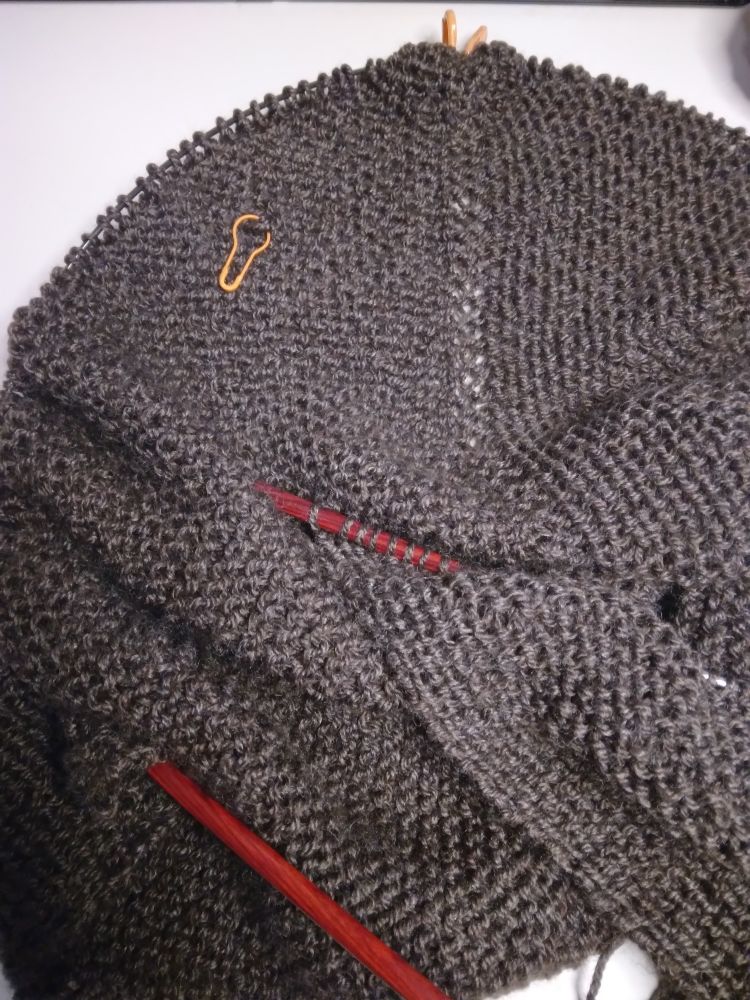 A piece of knit fabric made from a dark brown-grey yarn. A few inches under the live stitches, an orange stitch marker holds a loose stitch to keep it from unraveling. 