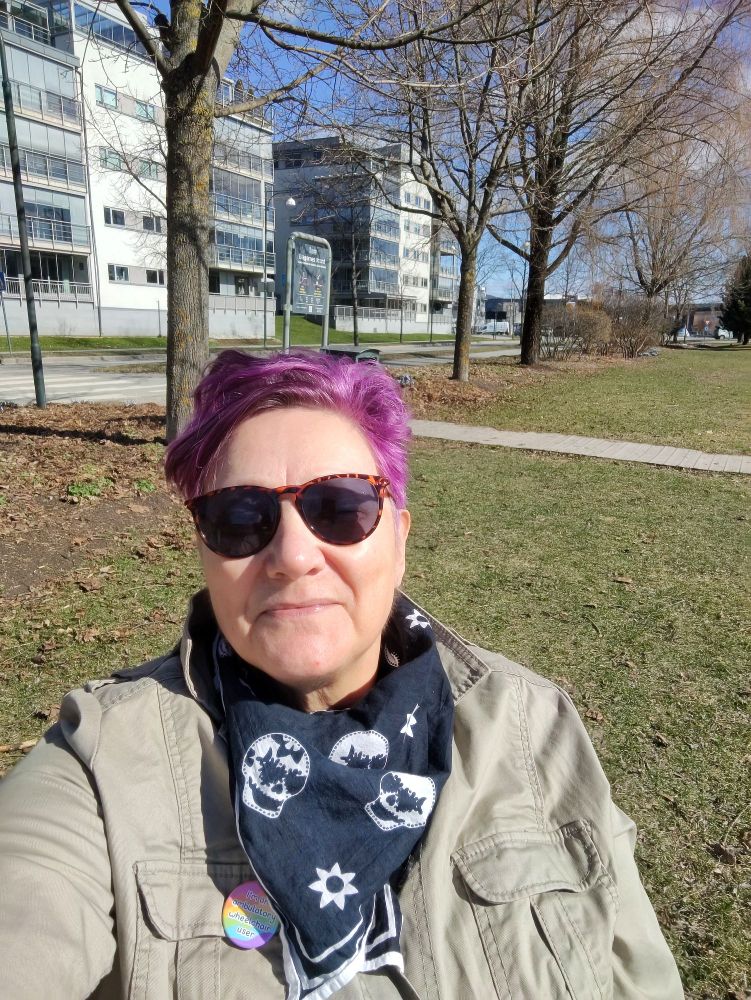 Jeg sitter på en begynnende grønn plen med trær og bygninger i bakgrunnen. Jeg har kort, knall rosa hår, mørke solbriller, svart skjerf med hvite hodeskaller og en beige jakke.