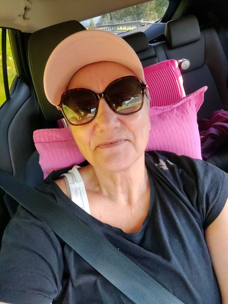 En middelaldrende kvinne sittende i passasjersetet på en bil, med sikkerhetsbelte på. En rosa caps dekker håret, jeg har store, mørke solbriller og en hvit singlet under sort t-shirt. 