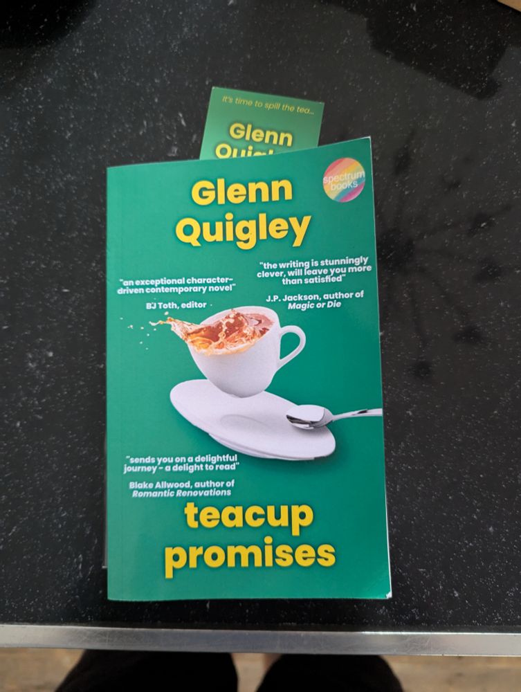 Front cover of Glenn Quigley's book Teacup Promises. A green background has a photo of a full teacup, saucer and teaspoon. The teacup looks like it has been knocked and rea is starting to splash.