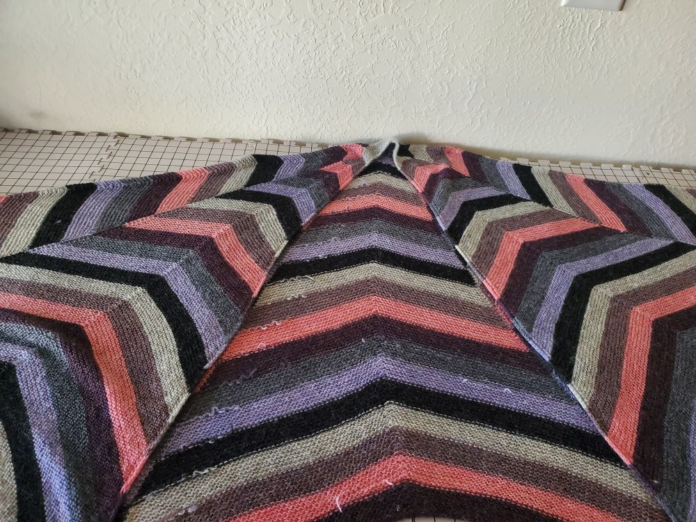 Very large shawl, that doesn't entirely fit in the frame.  The shawl is created using 7 colors in 7 wedges, with the stripe pattern shifting by one layer each wedge the first wedge.  Unfortunately, I'd couldn't take a picture with all of it visible, as I would need a bigger house and 2 more sets of blocking mats, so it's folded with a wedge and a half on top, each side, and one wrong side wedge in the middle, where several woven in ends can be seen.  The colors are silver, a heathered cool redish color, pink, deep purple leaning towards red, dark grey, lavender, and black.  