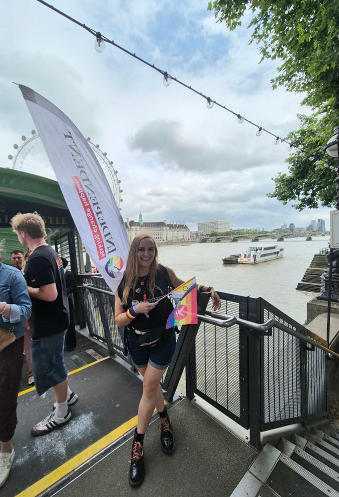 Sian smiling by the river at the end of Pride parade, holding an Independent flag