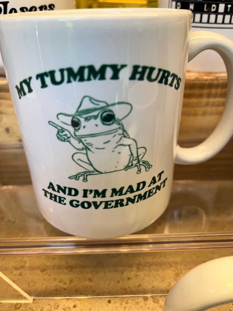 White mug with a picture of a frog wearing a fedora and green text that says “my tummy hurts and I’m mad at the government.”