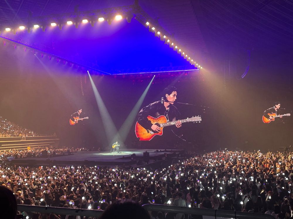 Suga of BTS in concert at the Singapore Indoor Stadium. He’s playing a guitar signed by the BTS members and seated alone in the middle of the stage. Around him are armys waving army bombs. 