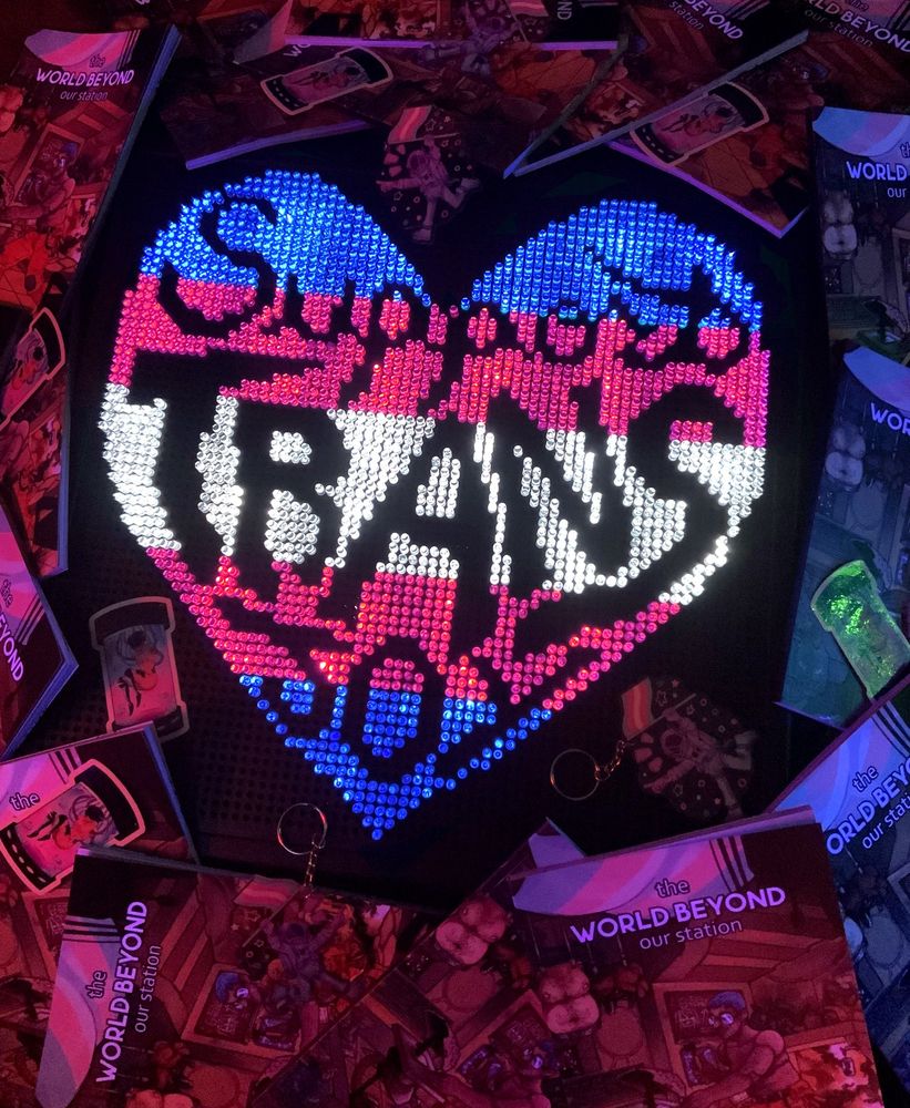 Lite brite art of a heart in trans pride colors with the text "support trans joy" on it. It's surrounded by copies of the World Beyond our Station sci-fi transmasc zine, plus some merch.