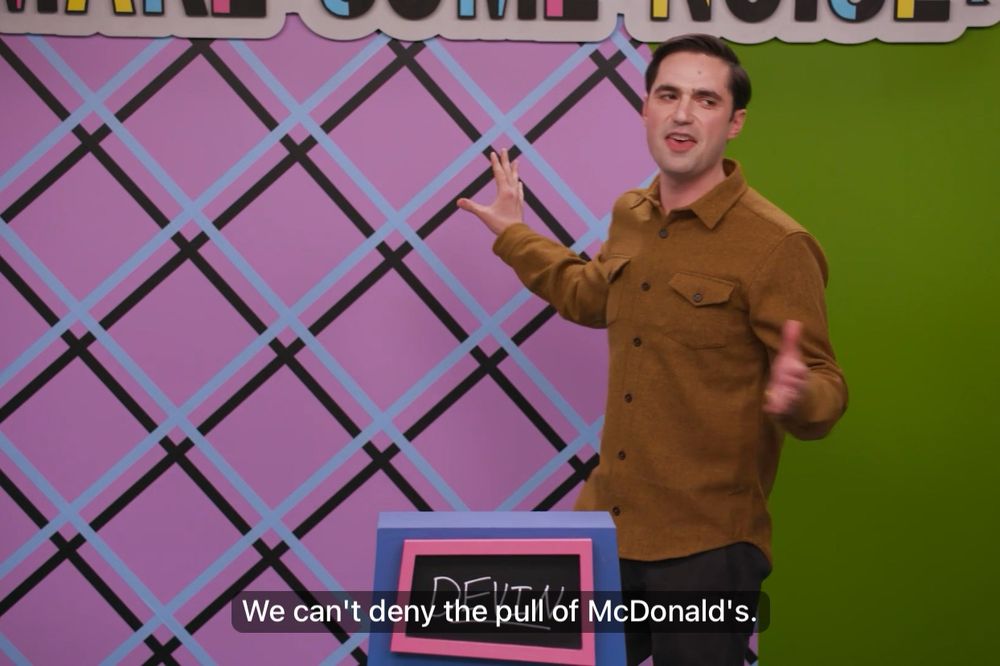 Devin on Make Some Noise on Dropout, wearing a brown button up shirt and black slacks, standing behind a pink and purple slanted podium with his own name on it written in chalk. Behind him is a green wall with pink on the wall to his right. He’s facing camera but looking off camera, arms outstretched. Caption reads “We can’t deny the pull of McDonald’s”