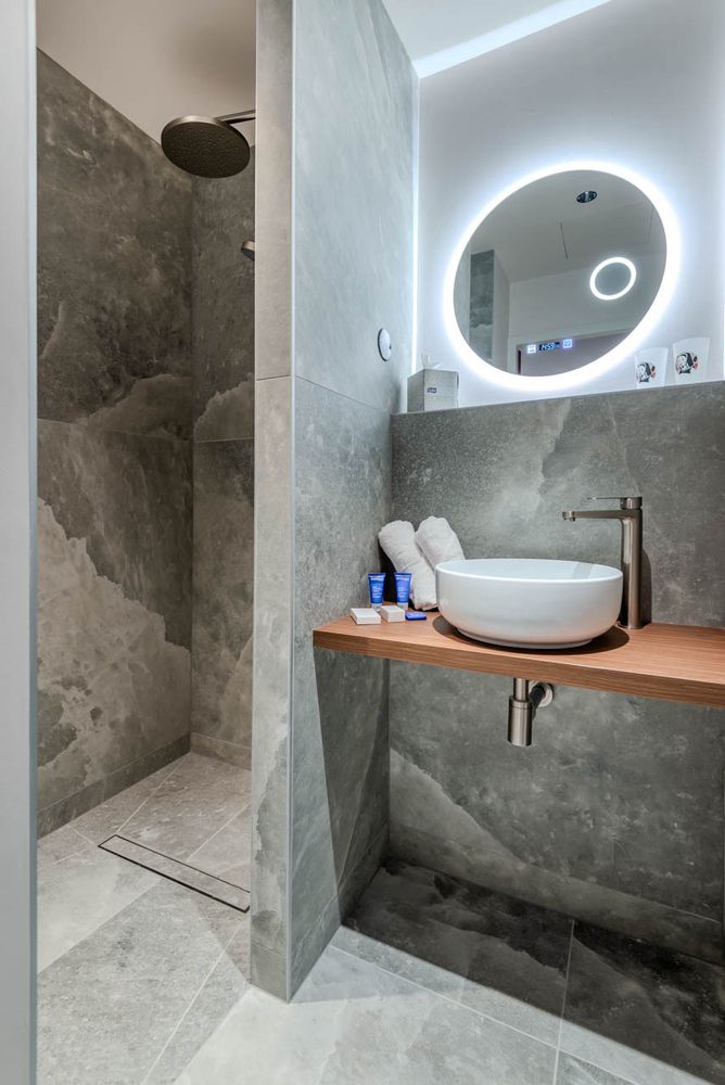 La salle de bain se compose d’une douche à l’italienne, en pierre grise, une paroi de cette même pierre sépare la partie lavabo qui est une vasque ronde blanche posée sur un plan en bois. Un miroir rond avec un néon de lumière derrière éclaire le tout.
Il y a des produits de beauté Phytomer et 2 serviettes de toilettes disposées en rond à côté de la vasque.