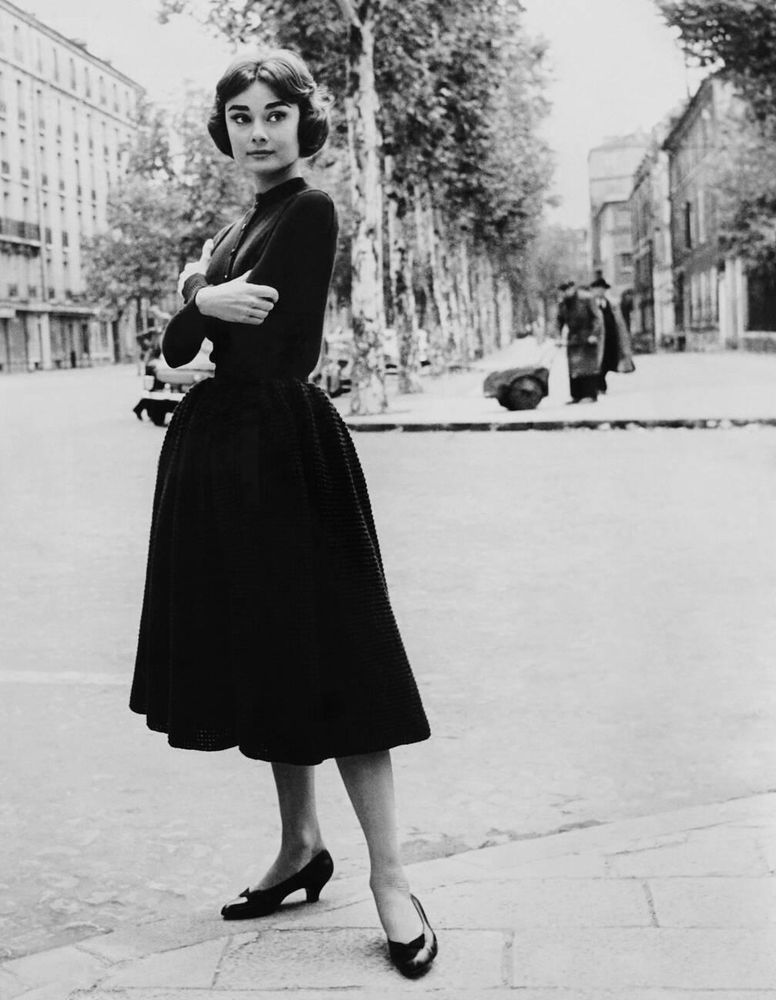 Actress Audrey Hepburn — in costume, dressed in an elegant dark-coloured gown designed by Hubert de Givenchy — captured in profile, face turned toward the camera, her arms crossed, while standing on the edge of a pavement, the street in the background replete with buildings, trees and passersby, in a full-length black-and-white shot taken in Paris during a break from location filming for Love in the Afternoon (1957)