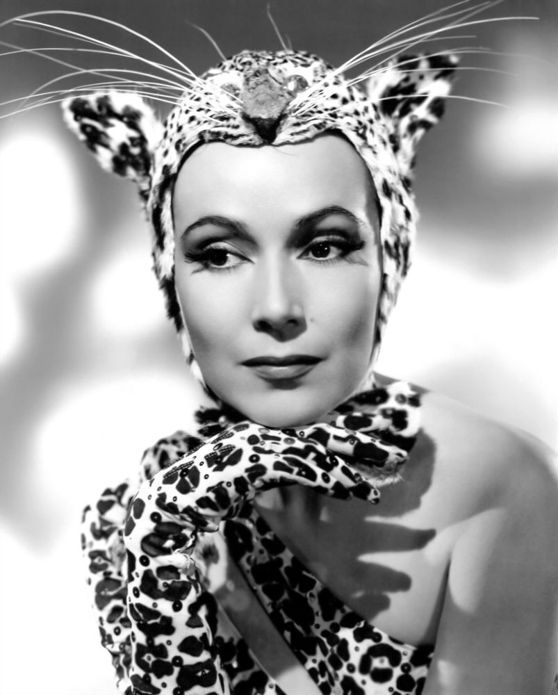 Actress Dolores del Río — in eccentric costume, dressed in a one shoulder leopard print bodysuit, gloves and leopard head headdress replete with ears and large whiskers — captured facing the camera while glancing to the right, her chin resting on her right hand, posed against a light-coloured background, in a black-and-white medium close-up shot for the film Journey Into Fear (1943)