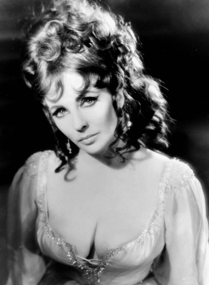 Actress Elizabeth Taylor — in costume, dressed in a light-coloured gown with front-laced corset and shimmering accoutrements — captured facing the camera while looking directly into the lens, her head tilted slightly to the right, posed against a black background, in a medium-length black-and-white shot for the film Doctor Faustus (1967)