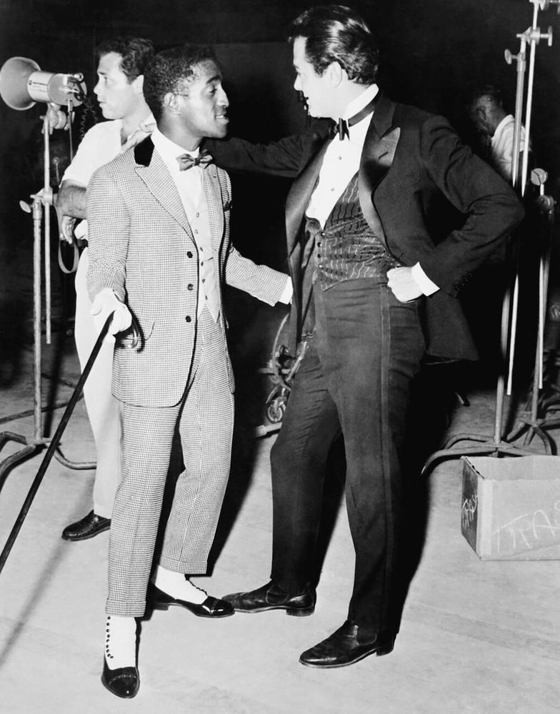 Singer, actor and dancer Sammy Davis Jr., and actor Tony Curtis — both in costume, Davis dressed in a light-coloured three-piece suit, bowtie and white spats, Curtis in a black three-piece tuxedo — captured facing each other while standing together in profile, several pieces of film and lighting equipment in the background, a crew member behind them, in a full-length black-and-white shot taken during a break from filming on the set of Some Like It Hot (1959)