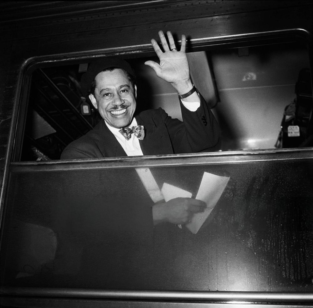 Musician and bandleader Cab Calloway — dressed in a black suit and light-coloured patterned bowtie — captured facing the camera while looking through the open window of a train carriage and smiling broadly, waving with his left hand and holding some papers in his right, in a medium-length black-and-white shot taken from the platform at a train station in Paris