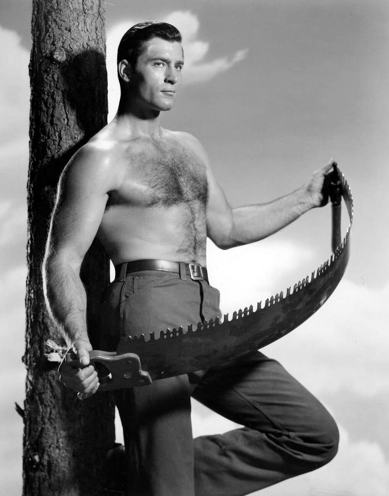 Actor Clint Walker — shirtless, dressed in dark-coloured trousers — captured standing in profile while leaning his back against a tree trunk, face turned slightly to the right as he looks into the distance, a long saw held between his hands, its blade bending, in a medium-length black-and-white shot for the television series Cheyenne