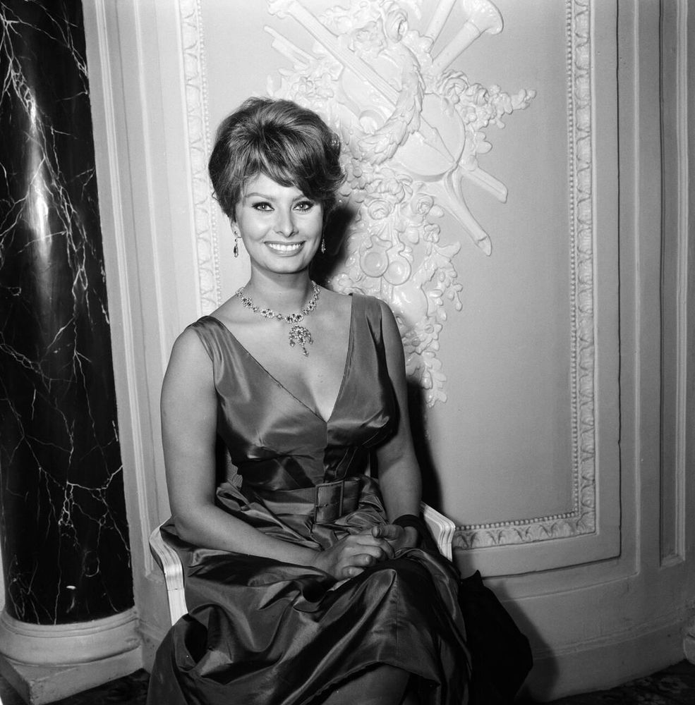 Actress Sophia Loren —dressed in a dark-coloured V-neck-shaped gown — captured facing the camera while sitting on an elegant chair, looking toward the camera and smiling broadly, her hands clasped together in her lap, in a medium-length black-and-white shot taken at the Ritz Hotel in London during a press conference to promote the film The Millionairess (1960)