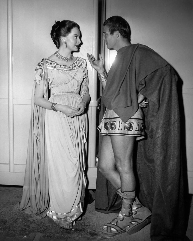 Actors Deborah Kerr and Marlon Brando — both in costume, dressed in Roman costume, Kerr wearing a white stola, Brando in a tunic and dark-coloured cloak — captured standing together, Kerr facing the camera, turning to look at Brando who leans against a wall in profile, a lit cigarette in his left hand, as they converse between takes, in a full-length black-and-white shot during a break from filming on the set of Julius Caesar (1953)