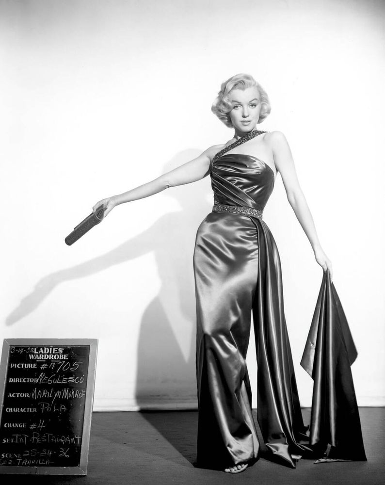 Actress and singer Marilyn Monroe — dressed in a dark-coloured one-shoulder evening gown with jewelled strap and belt details and long train designed by William Travilla — captured facing the camera while posing elegantly, holding a purse in her right hand while pointing toward a chalkboard etched with costume details, her left hand holding up the train of the gown, in a full-length black-and-white shot taken as part of a wardrobe test during production of the film How to Marry a Millionaire (1953)