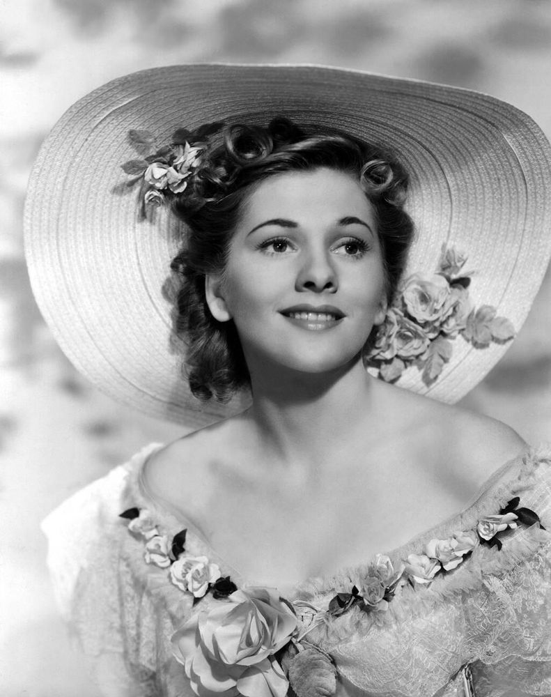 Actress Joan Fontaine — in costume, dressed in an elaborate light-coloured off-the-shoulder gown with flower-adorned bust, a replica of the titular character's dress, and wearing a wide-brimmed hat with flower accoutrements — captured facing the camera while smiling and glancing to the left, posing in front of a soft cloudy background, in a medium-length black-and-white shot for the film Rebecca (1940)