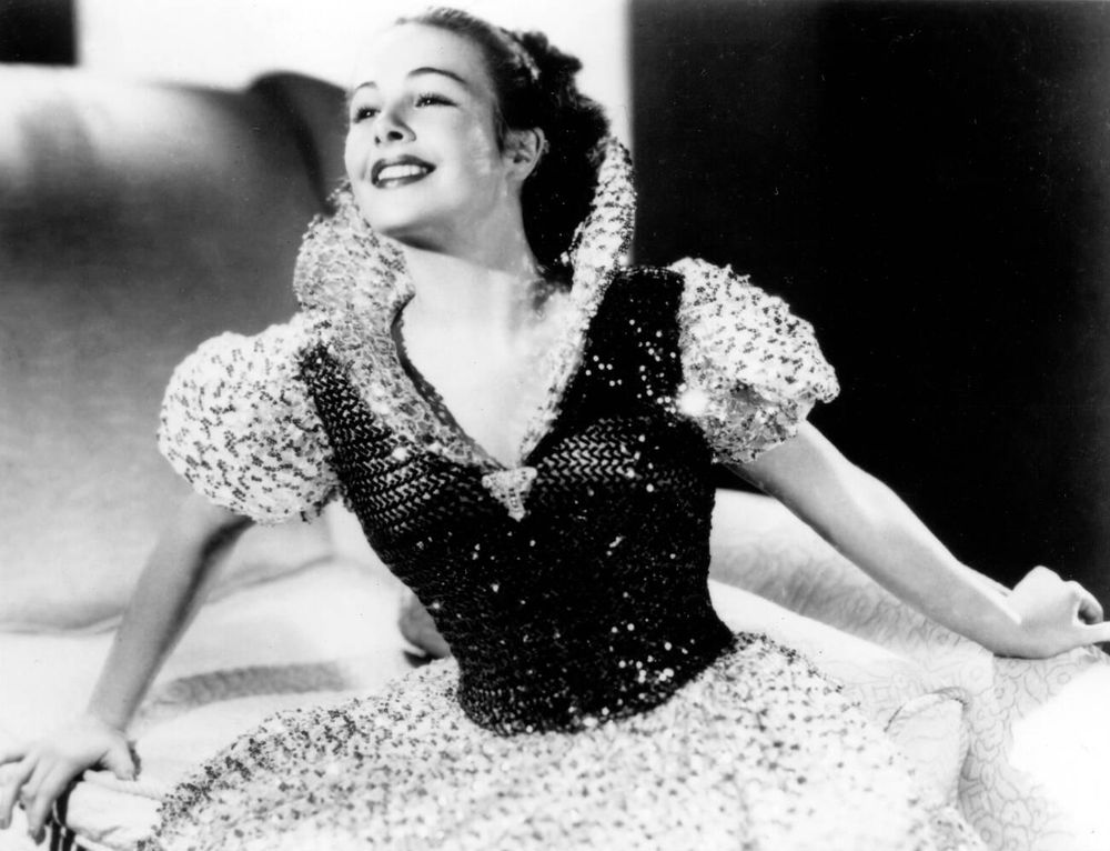 Dancer and actress Marge Champion — in Snow White costume, a beaded gown with dark-coloured bodice, light-coloured skirt, puffed sleeves and high collar — captured facing the camera while seated on an oversized chair, looking upward and smiling broadly, arms resting by her sides, in a medium-length black-and-white shot taken during live-action model reference tests for animators of the film Snow White and the Seven Dwarfs (1937)