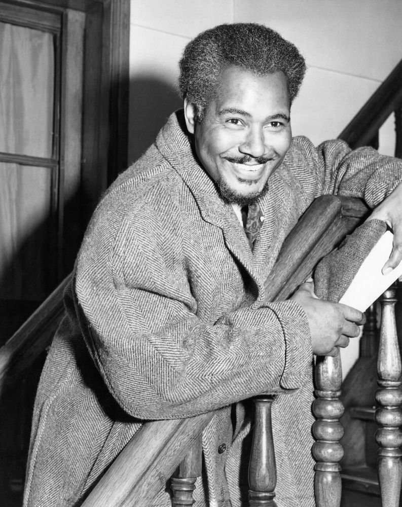 Actor Rex Ingram — dressed in a heavy dark-coloured coat — captured facing the camera while standing on a staircase and leaning on the handrail, looking off-camera, smiling broadly, in a medium-length black-and-white shot taken onboard the Italian ocean liner SS Rex while sailing to New York City shortly after Ingram completed production of the British film The Thief of Bagdad (1940)