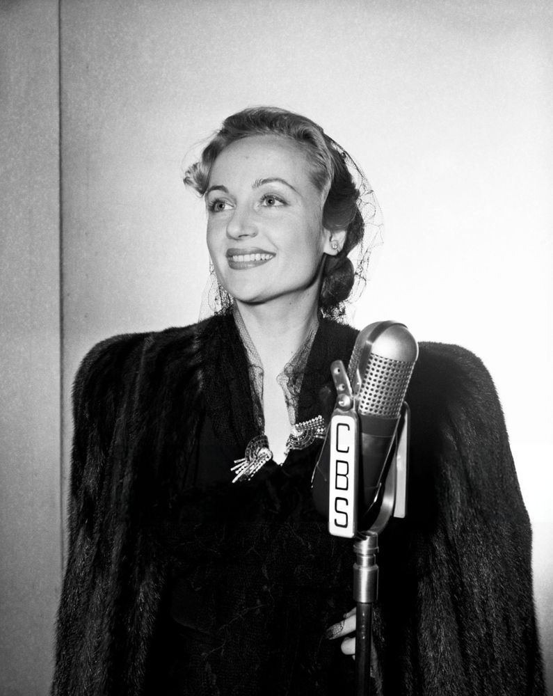 Actress Carole Lombard — dressed in a black dress and a veiled hat, a dark-coloured fur coat draped over her shoulders — captured facing the camera while glancing upward to the right, smiling broadly, a CBS-inscribed standing microphone positioned in front of her, in a medium-length black-and-white shot taken in the studio during recording of an episode of the radio series Silver Theater