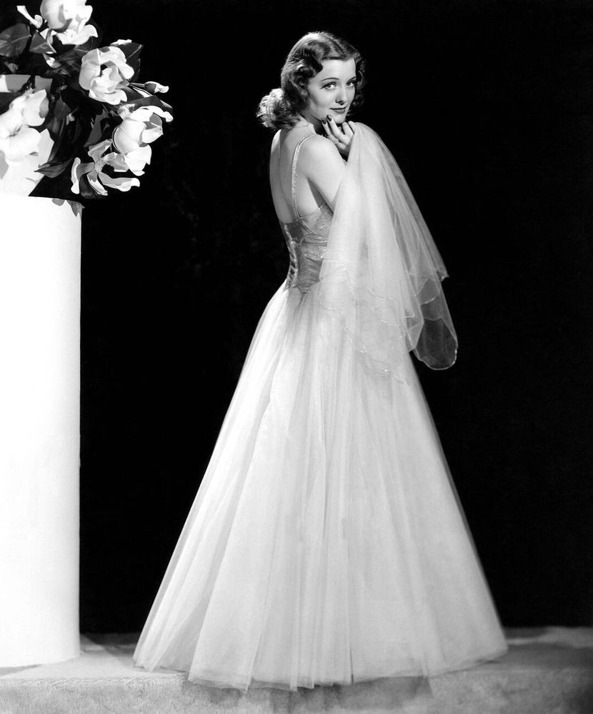 Actress Ellen Drew — in costume, dressed in a light-coloured gown with silver lamé bodice and voluminous net skirt designed by Edith Head — captured standing in profile, face turned to the camera, looking into the lens as she holds a large net handkerchief under her chin, a white column topped with a vase of flowers beside her, in a full-length black-and-white shot for the film The Mad Doctor (1941)