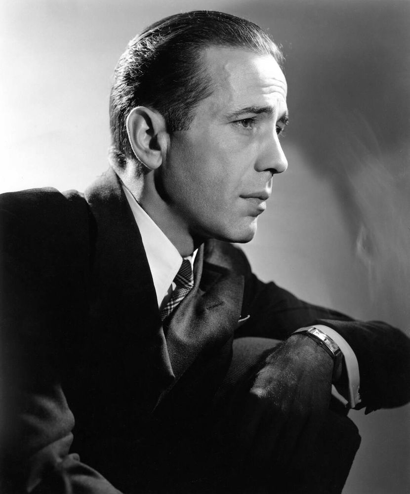 Actor Humphrey Bogart — dressed in a black suit and dark-coloured check patterned tie — captured in profile while seated on a dark chair, looking off-camera, his left arm resting on the chair arm, posed against a light-coloured background with mixed shading, in a medium-length black-and-white studio portrait taken during Bogart's tenure at Warner Bros.