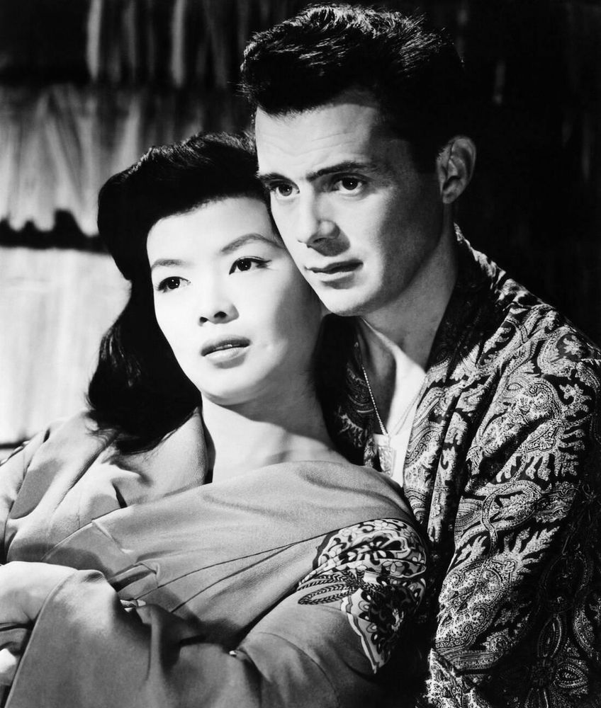 Actors Yoko Tani and Dirk Bogarde — both in costume, Tani dressed in a light-coloured robe, Bogarde in a black robe with elaborate exotic pattern — captured together, both almost in profile while seated and looking off-camera, Tani leaning back against Bogarde's chest, in a medium-length black-and-white shot for the film The Wind Cannot Read (1958)