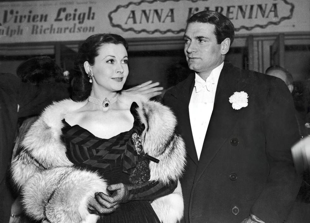 Actors Vivien Leigh and Laurence Olivier — Leigh dressed in a dark-coloured gown with striped bodice and sleeves and a light-coloured fur stole, Olivier in white tie and tails and a black coat — captured together, both facing the camera, Leigh looking to the left while Olivier glances in the opposite direction, the arm of an unseen man behind Leigh as a path through the crowd is created for the couple, in a medium-length black-and-white shot taken at the Leicester Square Theatre in London during the premiere of Anna Karenina (1948)