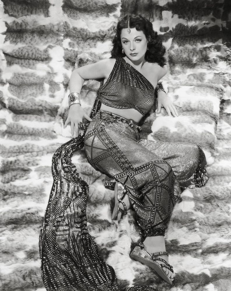 Actress Hedy Lamarr — in period costume, dressed in a shimmering exotic one-shoulder two-piece ensemble with train — captured facing the camera while lying on an extravagant plush surface, hands resting by her side as she looks upwards, in a full-length black-and-white shot taken from above for the film Samson and Delilah (1949)
