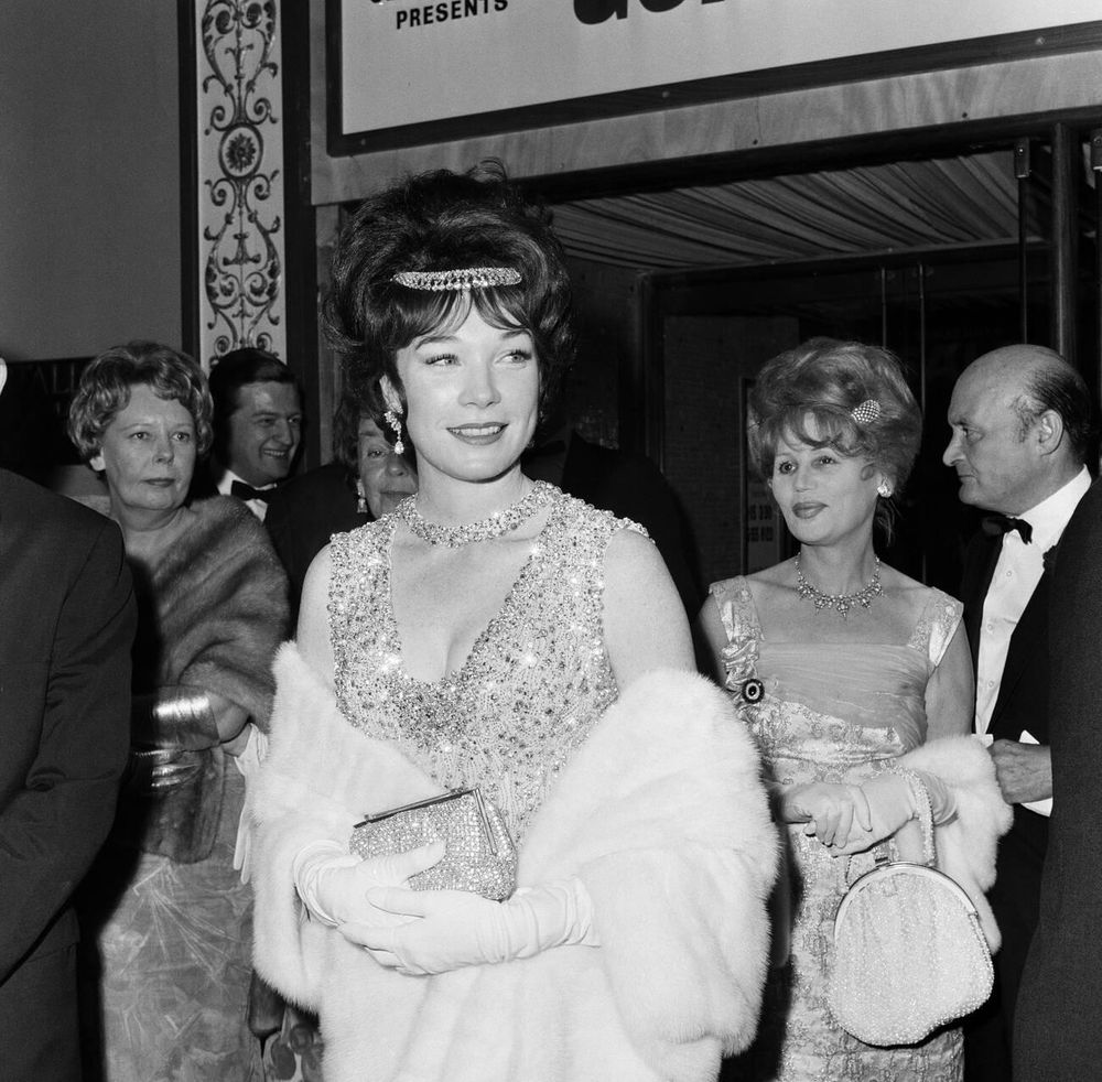 Actress Shirley MacLaine — dressed in a sparkling extravagantly beaded gown and white evening gloves, wrapped in a white fur stole and wearing a diamond tiara — captured facing the camera while standing, a shimmering purse in her hands, as she glances to the left, a number of well-dressed guests behind her, in a medium-length black-and-white shot taken outside the Carlton Cinema during the London premiere of What a Way to Go! (1964)