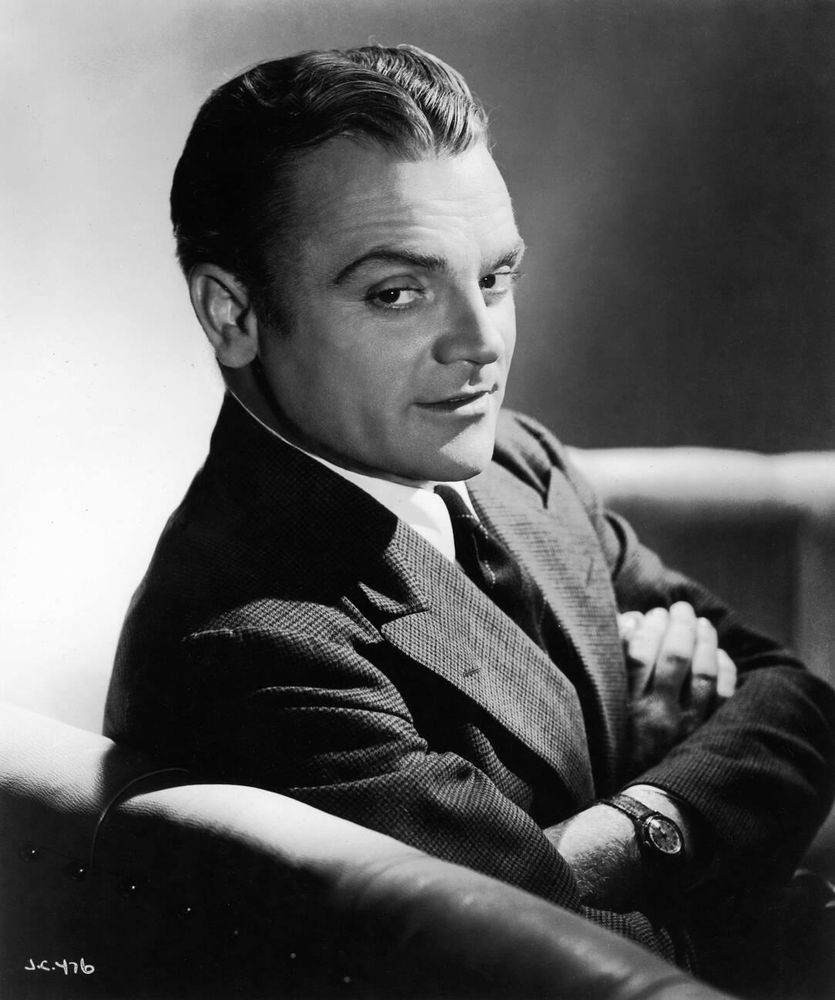 Actor and dancer James Cagney — dressed in a dark-coloured suit and tie — captured in profile while seated in an armchair, his face turned toward the camera as he raises his right eyebrow and smirks, arms folded, in a medium-length black-and-white shot taken by photographer George Hurrell