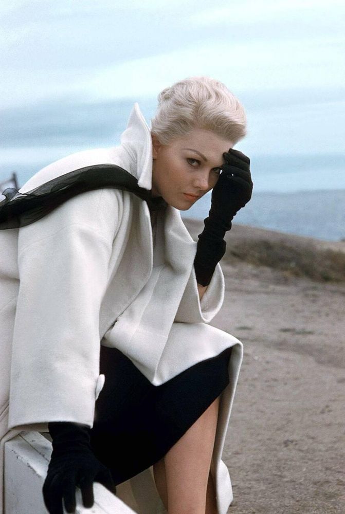 Actress Kim Novak — dressed in an elegant white coat, black scarf and long black gloves designed by Edith Head — captured in profile while seated, face turned toward the camera, her left hand resting against her forehead as she looks directly into the lens, San Francisco visible bay in the background, in a medium-length shot taken during a break from location filming for Vertigo (1958)