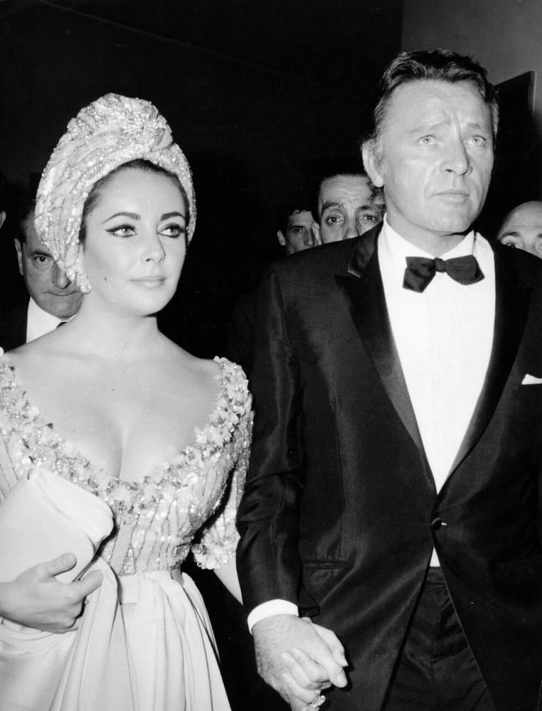 Actors Elizabeth Taylor and Richard Burton — Taylor dressed in a light-coloured off-the-shoulder gown with sparkling and matching turban, Burton in a black tuxedo — captured together, walking hand in hand as they arrive at the Silver Mask Awards, Taylor carrying a white purse, in a medium-length black-and-white shot taken at the Teatro Sistina in Rome