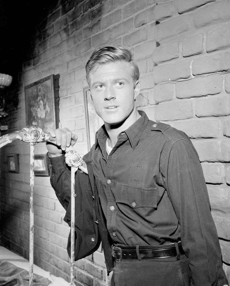 Actor Robert Redford — in costume, dressed in a dark-coloured shirt and trousers — captured facing the camera while standing in front of a bed, his hand resting on its metal headboard as he glances off-camera to the left, in a medium-length black-and-white shot for the television series The Twilight Zone