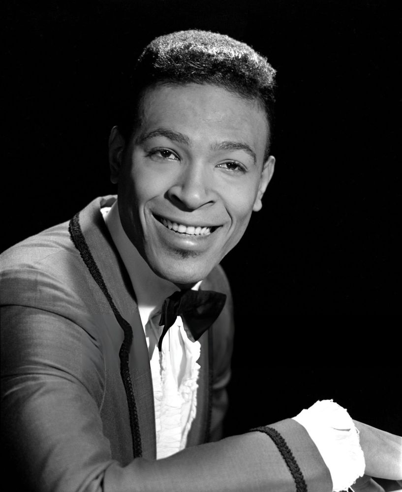 Singer and musician Marvin Gaye — dressed in a dark-coloured suit and black bowtie — captured in profile while seated, face turned toward the camera, smiling broadly, posed against a black background in a medium-length black-and-white studio portrait taken by photographer William "PoPsie" Randolph