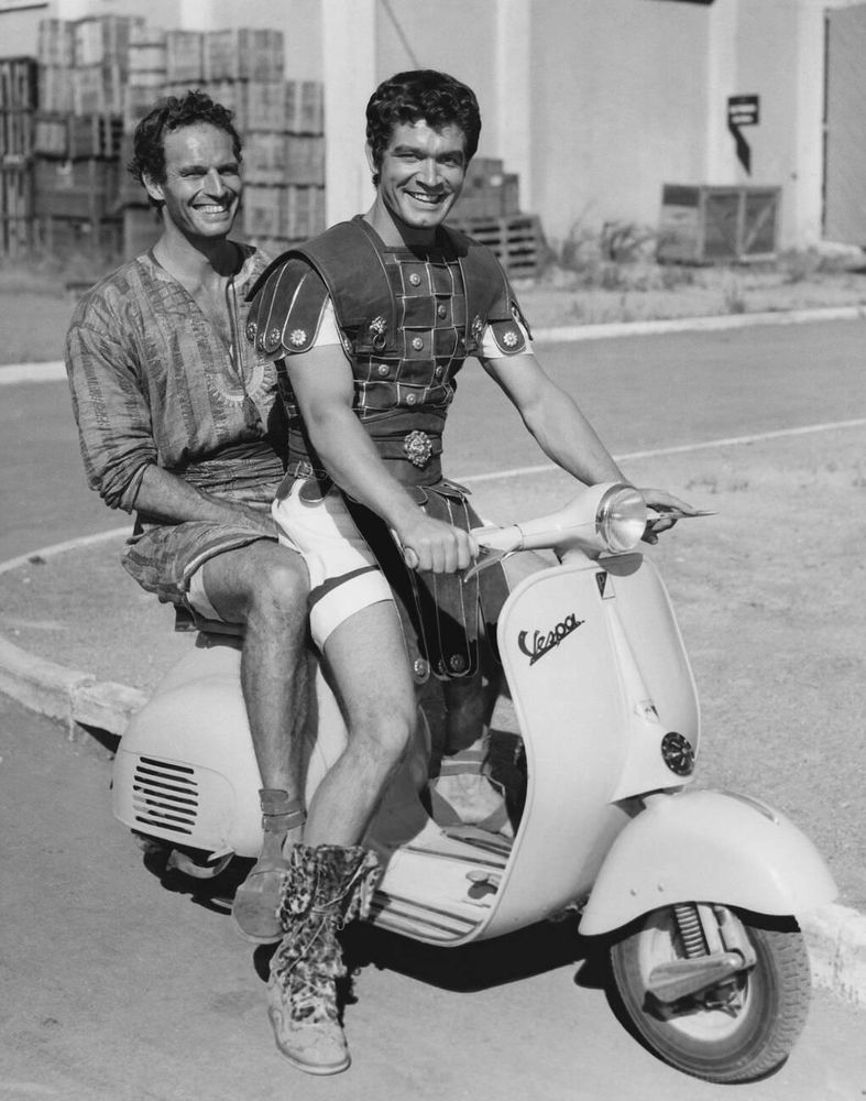 Actors Charlton Heston and Stephen Boyd — both dressed in period Roman costume — captured together almost in profile while seated on a light-coloured Vespa scooter, Boyd in front holding the handlebars as both turn to look at the camera, smiling broadly, in a full-length black-and-white shot taken on a street on the studio lot at Cinecittà during a break from filming Ben-Hur (1959)