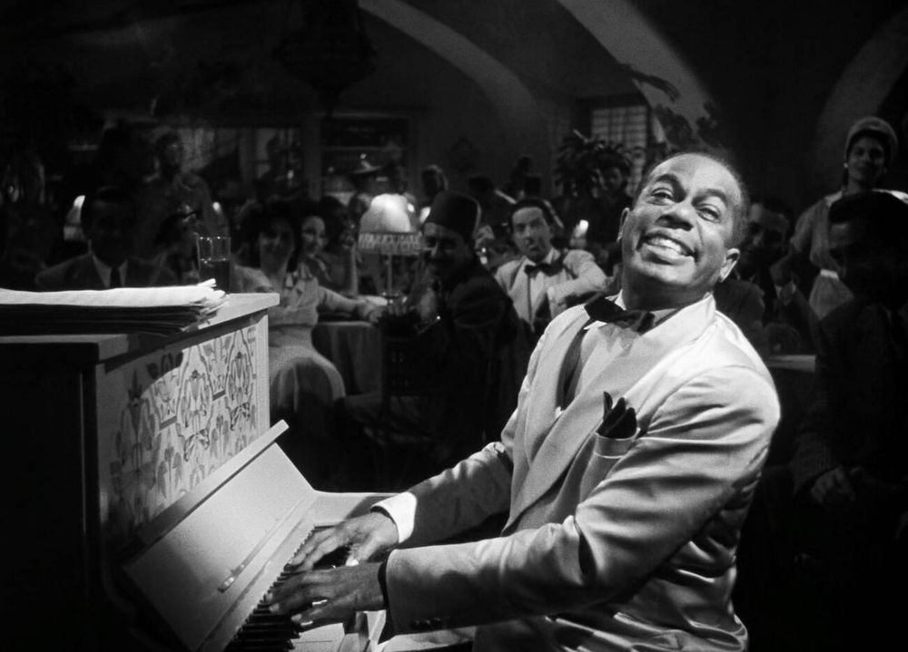 Actor and singer Dooley Wilson — in costume, dressed in a light-coloured tuxedo and black bowtie — captured sitting in profile at a piano, his fingers hovering above the keys as he turns his face toward the camera, head tilted to the left as he smiles broadly, a crowd seated behind him, in a medium-length black-and-white shot taken during filming of a scene at the fictional Rick's Café Américain in the film Casablanca (1942)
